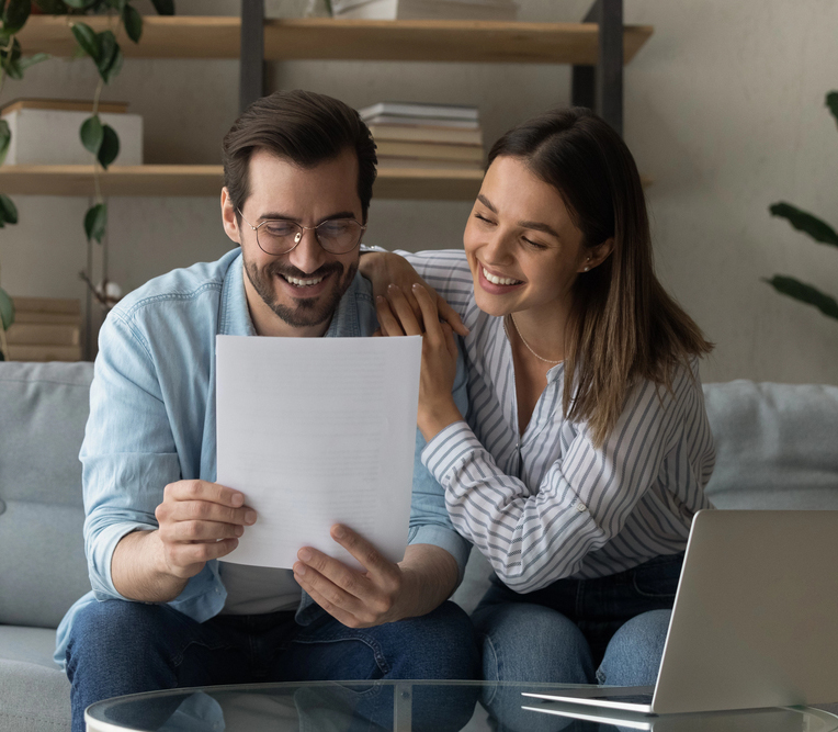 Happy young Caucasian couple feel overjoyed read good banking paper notice. Smiling millennial man and woman spouses sit on couch at home get pleasant news message in paperwork letter correspondence.